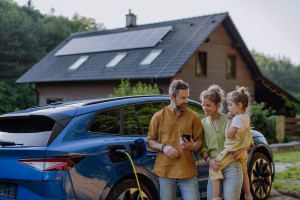 Familie an einem Elektroauto schaut aufs Handy vor einem Haus mit PV-Anlage auf dem Dach